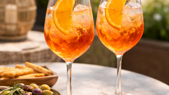 Two orange cocktails with orange slices and rosemary on a sunny outdoor table, plus a bowl of olives and breadsticks in the scene.