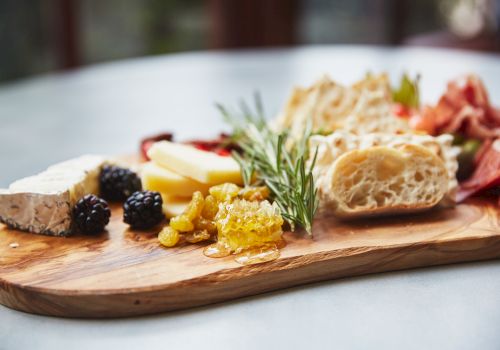 A cheese board with assorted items: cheeses, bread, olives, jam/honey, herbs, and cured meats on a wooden serving board.
