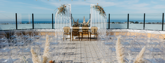 A wooden path leads to a deck on a sunny beach, with clear blue water, sandy shore, and potted plants along the walkway.