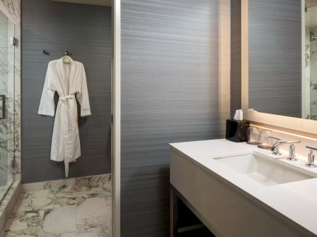 A modern bathroom with a white bathrobe hanging on a gray wall, a glass shower, marble floor, and a long white sink with a mirror and tidy counter.