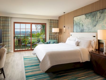 A bright hotel room with a large bed, nightstands, lamps, and a wall map; sliding glass door opens to a balcony with a view of greenery.
