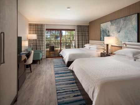 A hotel room with two queen beds, a desk, flat-screen TV, lamps, a blue rug, and a balcony view through glass doors.