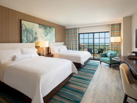 A hotel room with two queen beds, white linens, a large window balcony, a teal-striped rug, a seating area, and a desk with a chair.