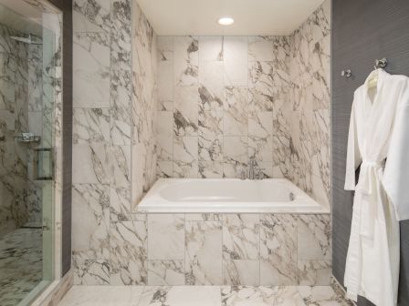 A luxurious marble bathroom with a built-in tub, glass shower on the left, and a white bathrobe hanging on a hook to the right.
