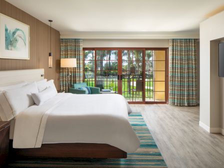 A bright hotel room with two queen beds, a seating area, and a large glass door opening to a garden patio; calming, modern decor.