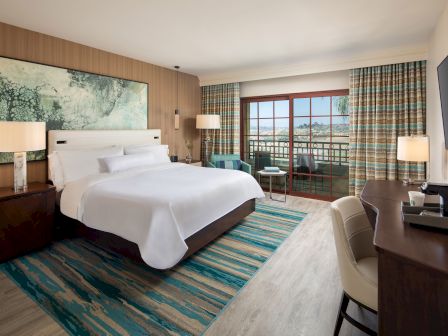 A hotel room with a large bed, wooden headboard, two nightstands, lamps, a desk with a chair, a rug, and sliding glass doors opening to a balcony with a scenic view.