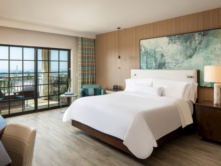 A modern hotel room with a large bed, wooden accents, a balcony patio, and ocean view outside, plus bedside lamps and artwork.