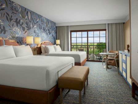 Two large beds with white linens in a modern hotel room, a patterned blue accent wall, a balcony view, and a seating area by a window.