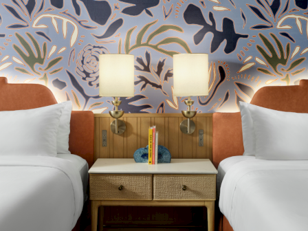 Two neatly made twin beds with white linens flank a wooden nightstand, two lamps, and bold botanical wallpaper behind.