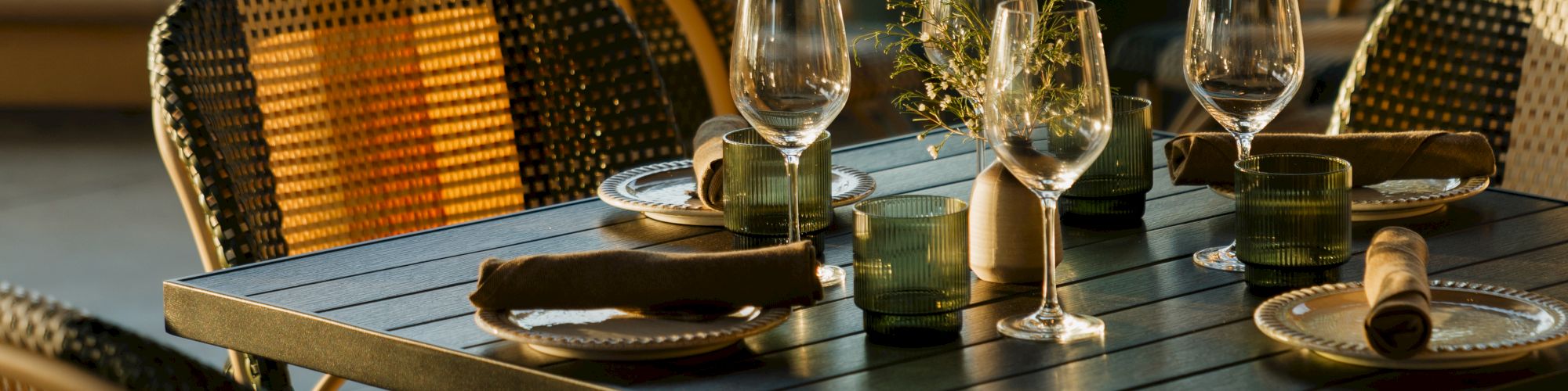 A stylish outdoor dining setup with a square black table, wicker chairs, wine glasses, and neatly arranged tableware by a poolside scene.