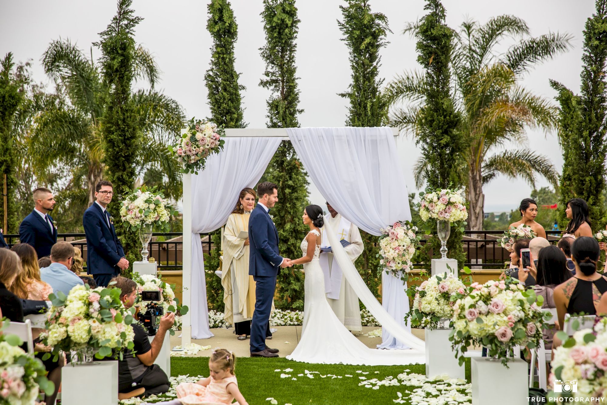 A wedding ceremony outdoors with a couple exchanging vows under a white draped arch, guests seated on either side, lush greenery and floral decor.