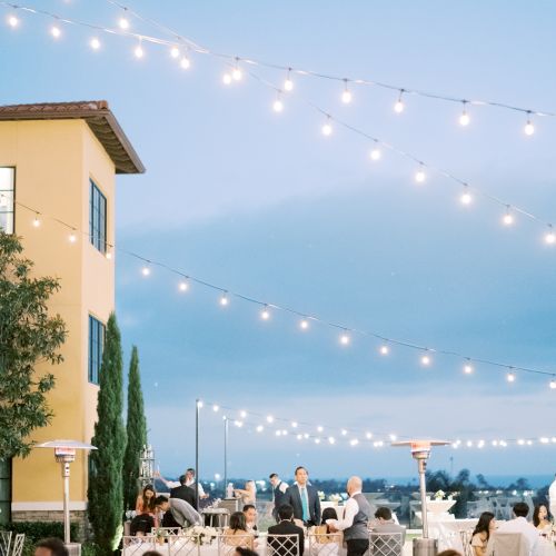 Outdoor evening wedding reception with string lights, guests seated at tables on a terrace, soft blue sky, and a yellow building in the foreground.