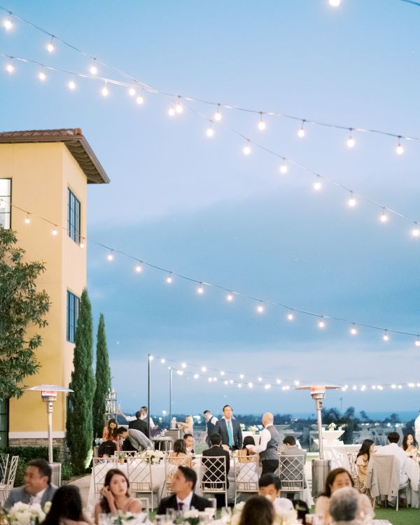 Outdoor evening wedding reception with string lights, guests seated at tables on a terrace, soft blue sky, and a yellow building in the foreground.
