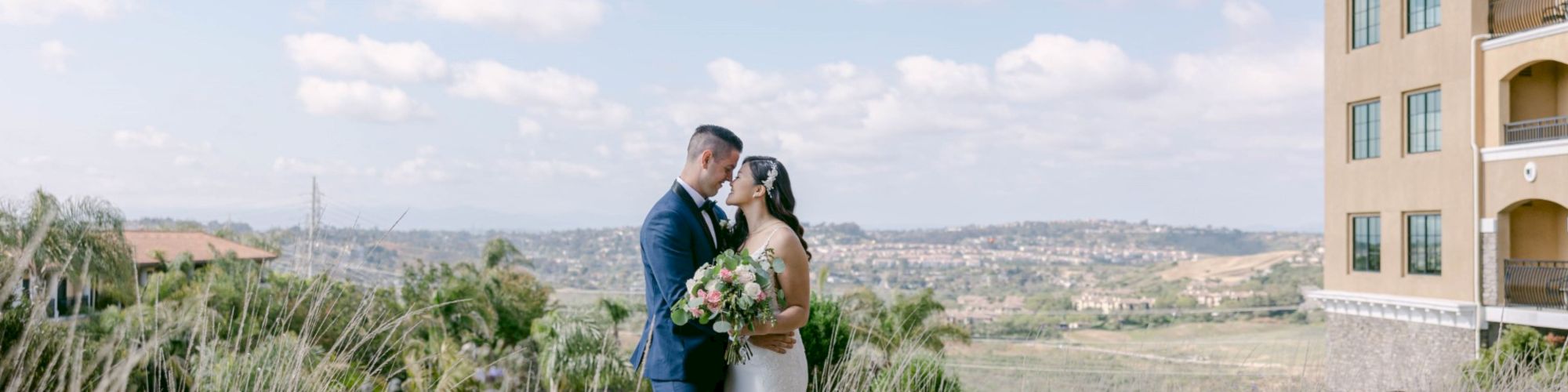 A newlywed couple in wedding attire share a kiss in a field of wildflowers, with a modern building in the background under a sunny sky.