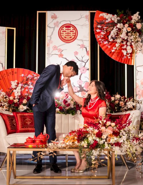 A couple on a decorated stage, exchanging a kiss or warm gesture, surrounded by red umbrellas, flowers, and ceremonial decor.