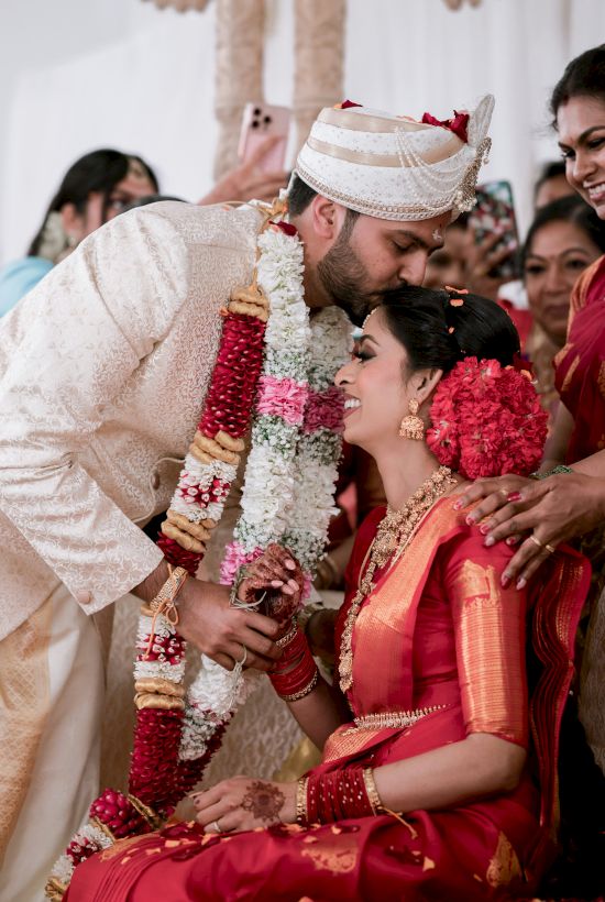 A groom bows to a bride in a traditional wedding, exchanging garlands amid joyous onlookers, vibrant red attire, and festive decor.