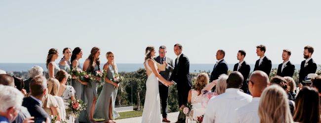A wedding ceremony outdoors: couple re-joins hands as officiant and bridal party stand nearby, guests seated and watching against a scenic backdrop.