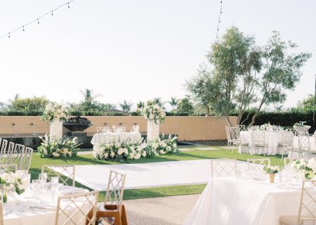An outdoor wedding setup with decorated tables, white flowers, string lights, and elegant chairs on a sunny lawn, ready for guests to arrive.