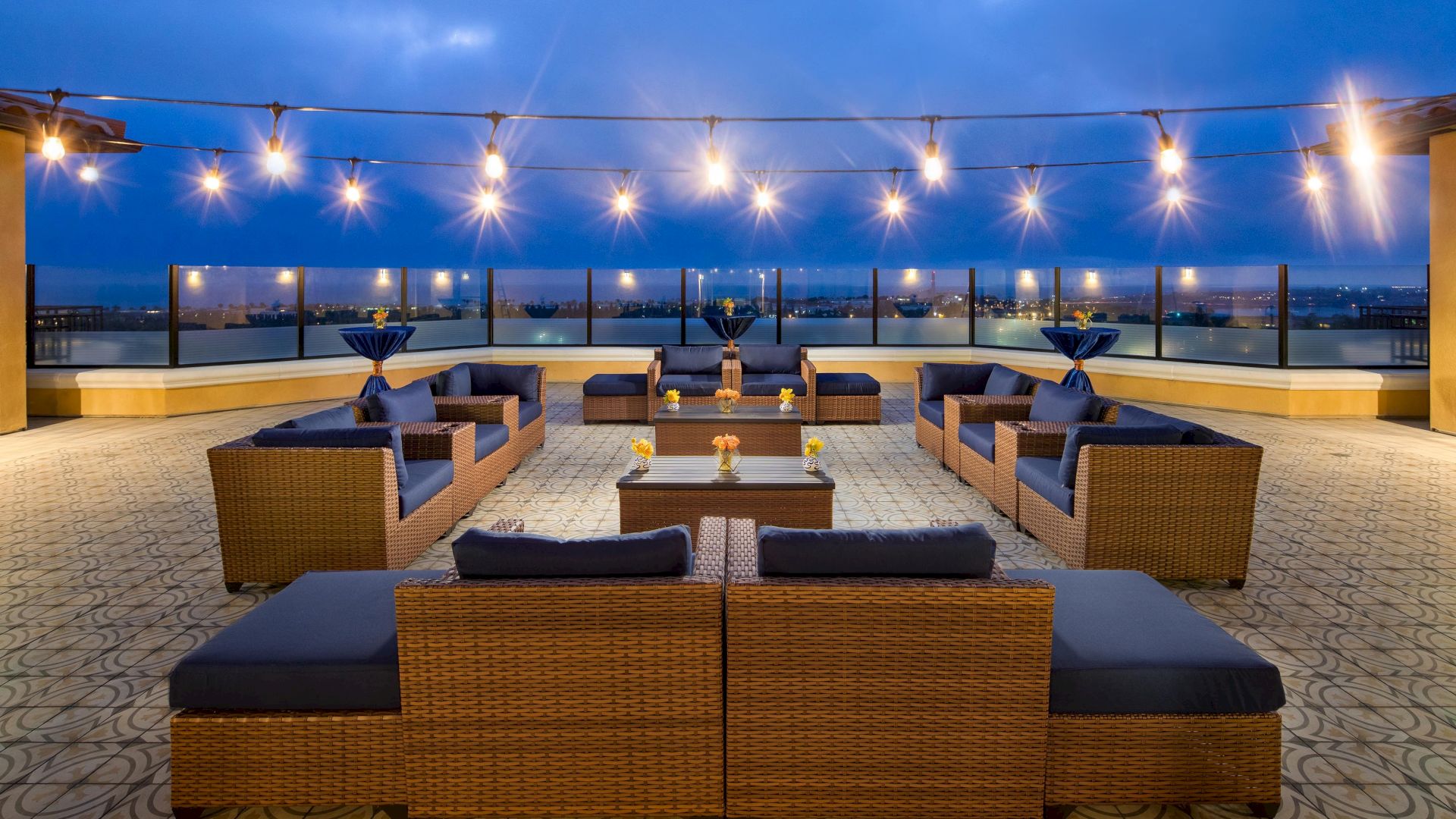 Hotel rooftop lounge with wicker sofas, low tables, string lights, and a city skyline in the distance, set for a chic evening gathering.