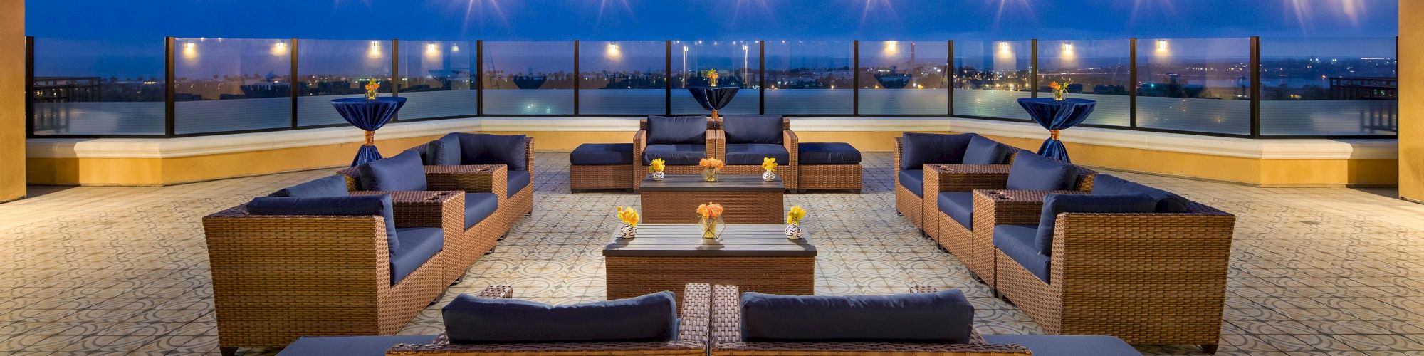 Hotel rooftop lounge with wicker sofas, low tables, string lights, and a city skyline in the distance, set for a chic evening gathering.
