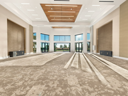 A bright, spacious lobby with tall windows, beige walls, and a wooden ceiling panel; sunlight streams in creating long shadows across a carpeted floor.