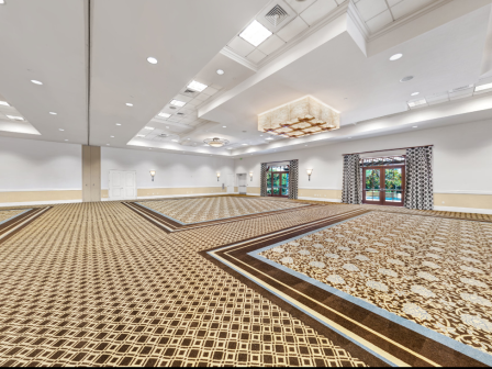 A large, empty banquet hall with patterned carpet, white walls, high ceilings, chandeliers, and multiple doors along the far wall.