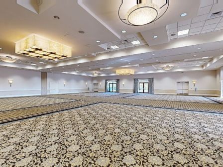 A large, empty conference ballroom with patterned carpet, high ceilings, chandeliers, and far double doors, ready for an event.