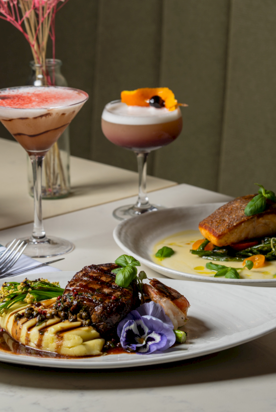 A fine dining spread: plated steak with veggies, a creamy sauce, a breaded side, and two elegant cocktails in stemware.