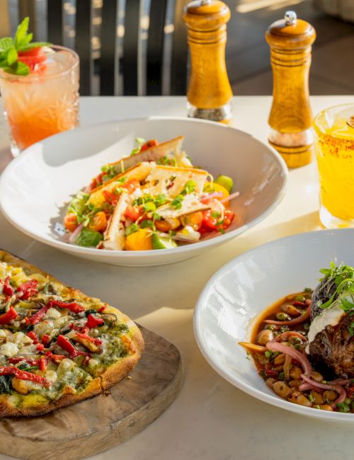 A sunny patio table with two pizzas, a salad bowl, steak dinner, and orange drinks with pepper mills.