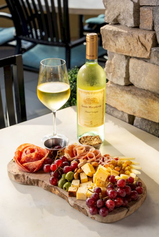 A charcuterie board with grapes, cheese, salami, and a glass of white wine beside a bottle on a sunny patio.