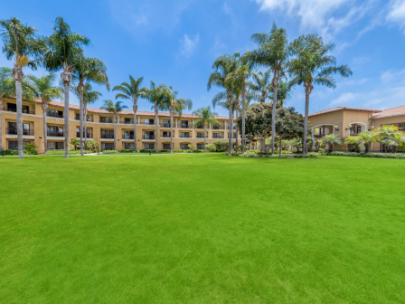 A sunny resort courtyard with a bright green lawn, palm trees, and low-rise beige buildings surrounding a wide open space, under a blue sky.