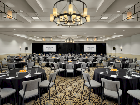 A large banquet hall set for a conference with round black-draped tables, gray chairs, elegant chandeliers, and a stage with screens at the front.