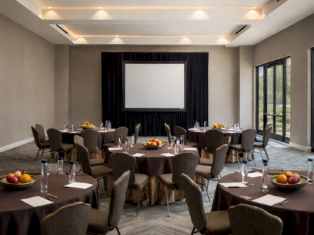 A modern meeting room set for a presentation, with round tables, cushioned chairs, glassware, and a projector screen.