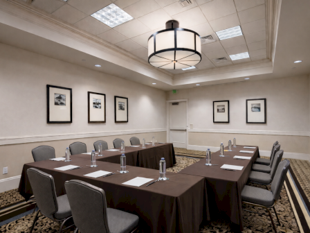 A conference room setup with a U-shaped table, brown tablecloths, gray chairs, water bottles, notepads, and framed pictures on beige walls.