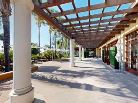 A sunny outdoor shopping arcade with columns, a covered walkway, palm trees, and storefronts along the path.