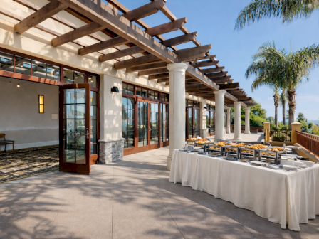 Sunlit resort terrace with wooden pergola, white columns, and a catered buffet of chafing dishes and pastries lined up.