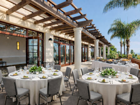 Outdoor restaurant patio with round tables set for al fresco dining under a wooden pergola, palm trees, and ocean view. Top it at 140 characters, always ending the sentence.