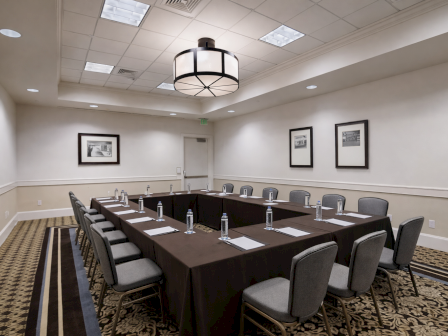 A formal conference room with a U-shaped table, gray chairs, water bottles, notepads, framed pictures on the walls, and a modern ceiling light.