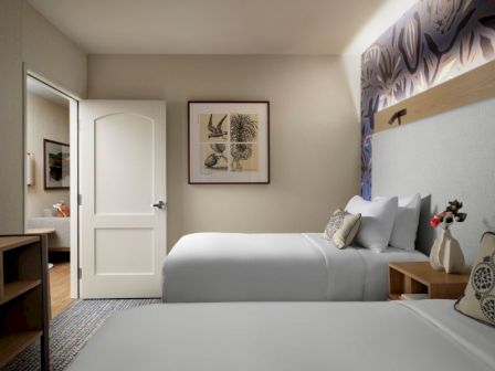 A modern hotel room with two neatly made beds, soft lighting, a framed decor piece, and a desk area near an open door to the bathroom.