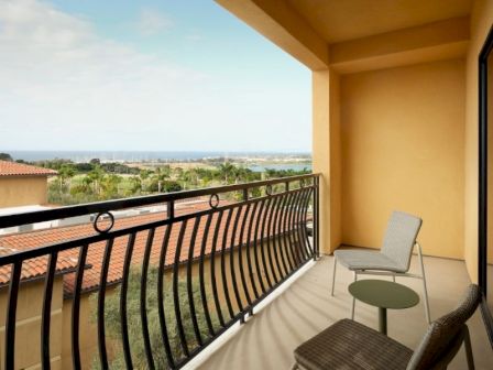 A balcony with beige walls, a small table and two chairs overlooks a sunny landscape with rooftops and distant trees toward the horizon.