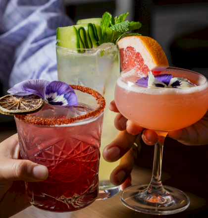 Two hands clink glasses of colorful cocktails&mdash;garnished with mint, citrus slices, and edible flowers.