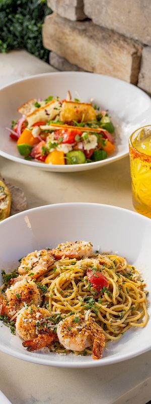 A sunny outdoor meal: spaghetti with shrimp, a colorful salad, a pizza slice, and a lemon drink.