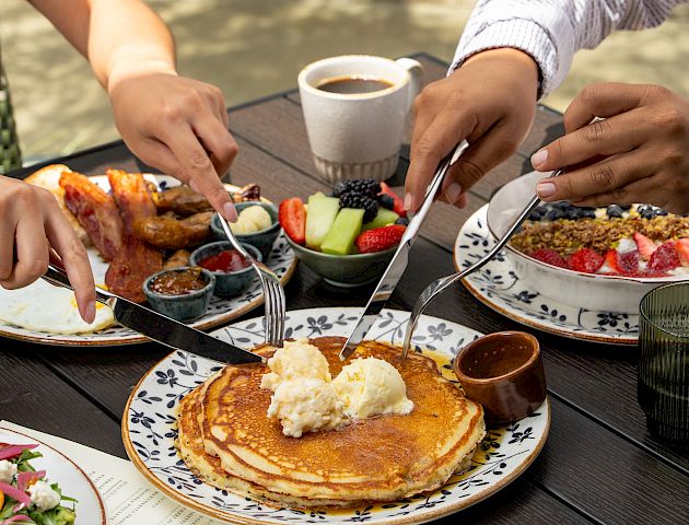 People enjoy a sunny outdoor brunch at a wooden table with pancakes, fresh fruit, and coffee by a riverside.