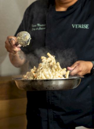 A person in a hoodie serves a bubbling, cheesy dish with noodles from a large pot, holding a spoonful for the camera.