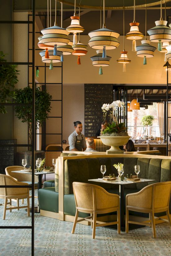 A chic restaurant interior with green chairs, glassware, and colorful hanging pendant lights over a bar where staff serve guests.