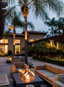 A modern outdoor lounge with beige sectional seating surrounding a central fire pit, palm trees, and a lit house backdrop at dusk.