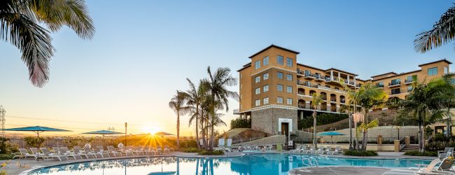 A resort-style swimming pool area at sunset with palm trees and a multi-story hotel in the background, calm and inviting atmosphere.