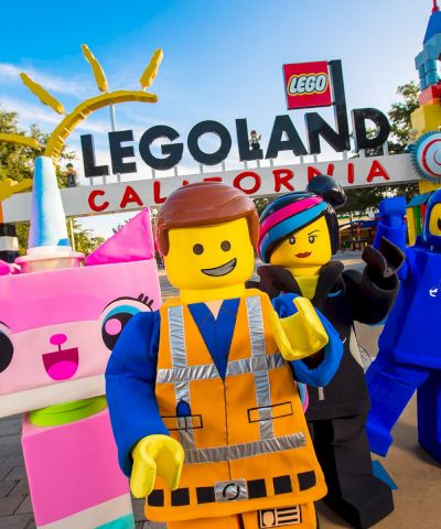 A group of LEGO characters posing in front of Legoland California, with a bright blue sky and colorful structures in the background. (140 chars max, ending with a period)
