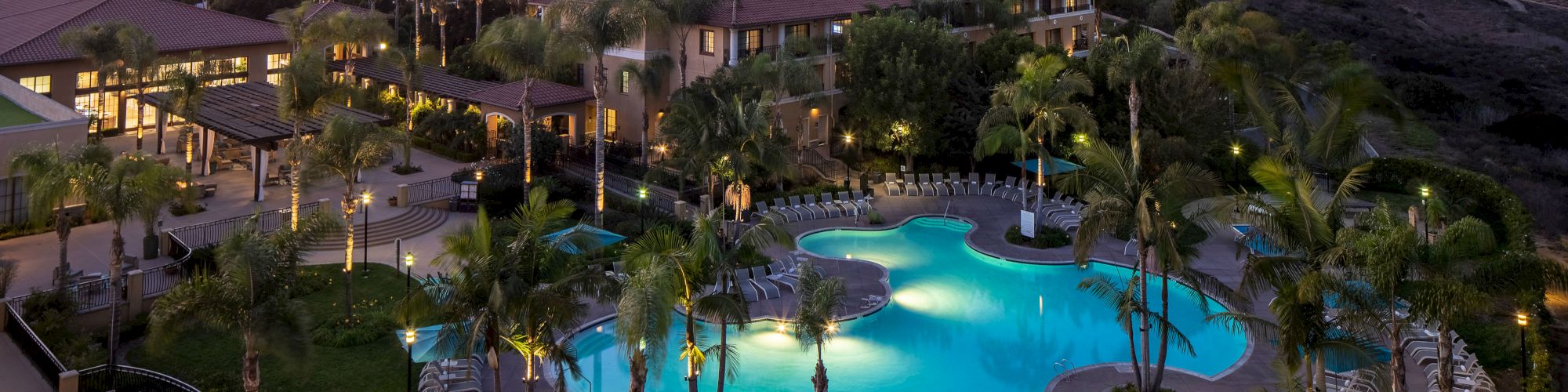 An aerial view of a resort with two illuminated pools, palm trees, lounge chairs, and surrounding low-rise buildings at sunset over a coastal landscape, serene.