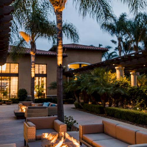 A modern outdoor lounge area with beige sofas, a built-in fire pit, palm trees, and warm lighting by a stylish house at dusk.